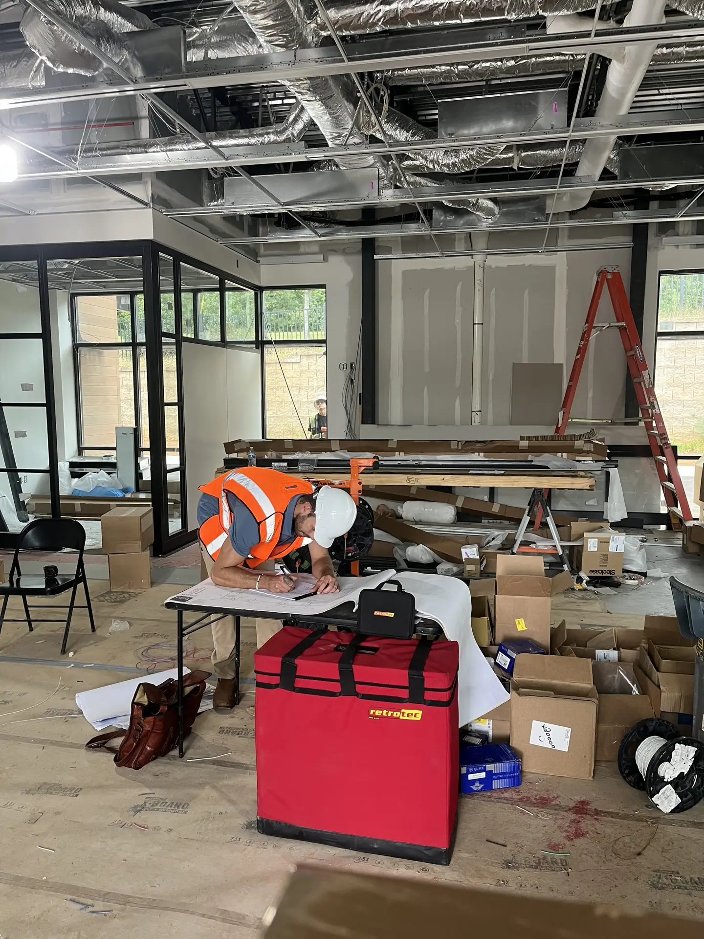 Construction worker in orange vest and hard hat reviewing blueprints at a job site with exposed ductwork overhead and a
